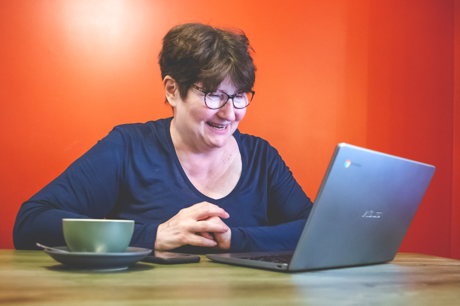 Simone Clarkin of A Life Less Ordinary Coaching sits at a bench desk looking at a laptop computer. A mobile phone and a coffee cup are beside her.