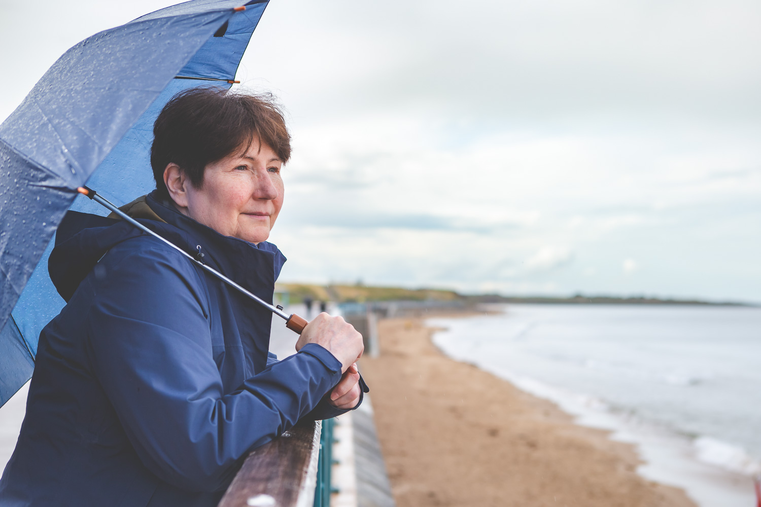 Simone Clarkin of allo coaching is looking out towards the sea wearing a dark blue jacket and holding an blue umbrella. In the background a promenade and beach can be seen.