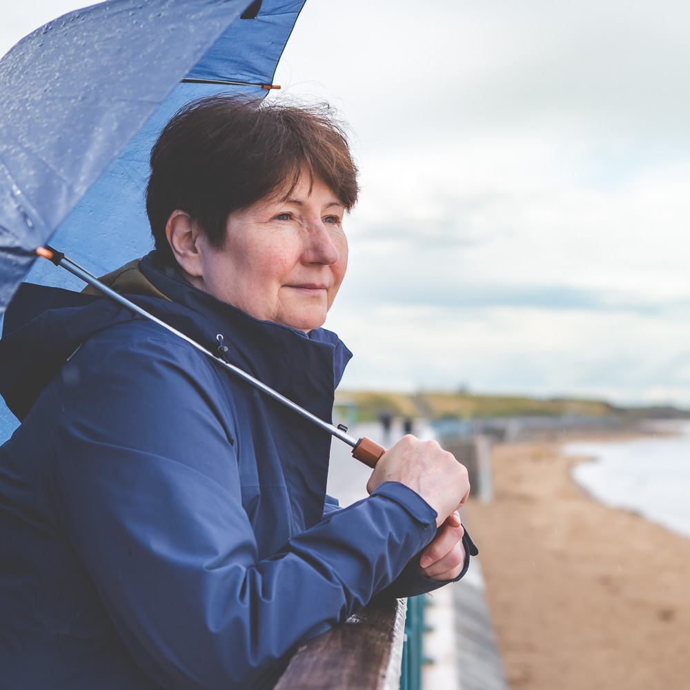 Simone Clarkin of allo coaching is looking out towards the sea wearing a dark blue jacket and holding an blue umbrella. In the background a promenade and beach can be seen.