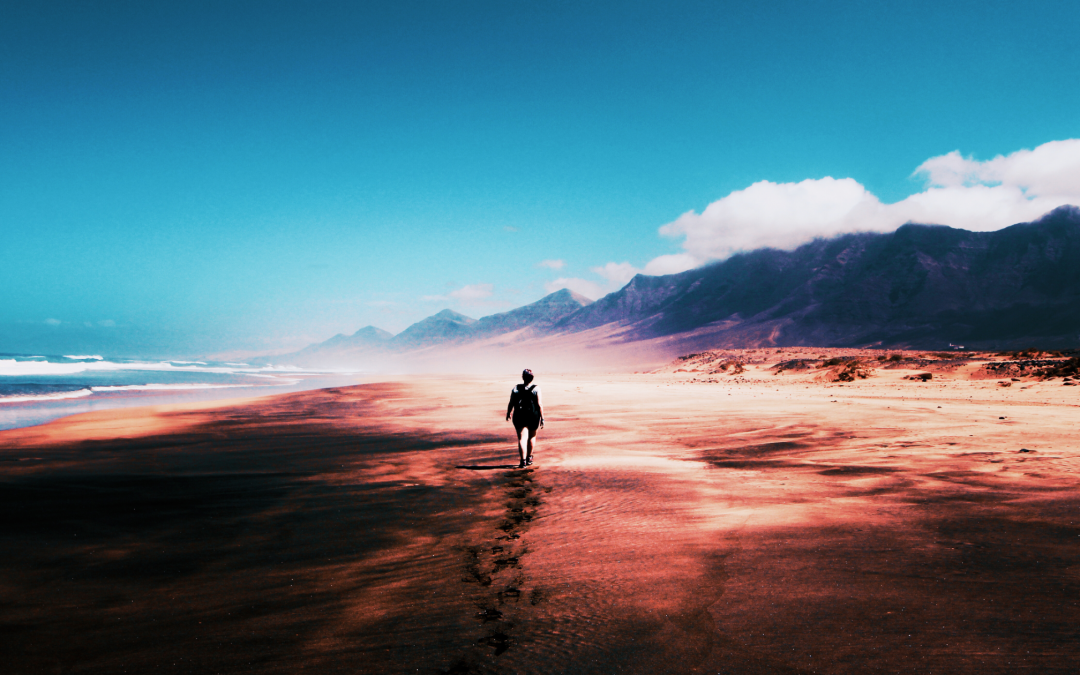 A solitary figure walks the shoreline, dwarfed by the immensity of the ocean and the sky. The deep blue expanse reflects the clear summer day, broken only by a handful of fluffy white clouds. Majestic mountains stand sentinel on the far right, their peaks shrouded in a soft mist. The rhythmic crash of waves is the only sound that breaks the silence, a constant reminder of the power and peace of nature.