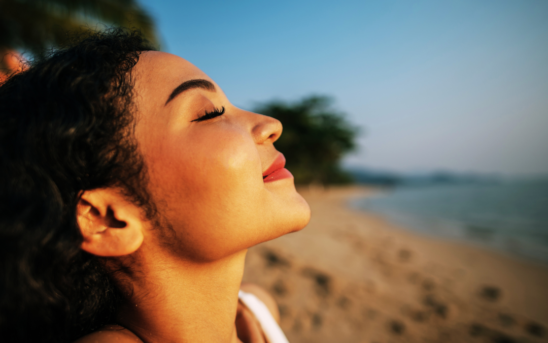 A relaxed young woman with long dark hair stands on a beach. Her face is tilted upwards, eyes closed in a smile, soaking up the sun. The background is blurred, but suggests a coastal setting.
