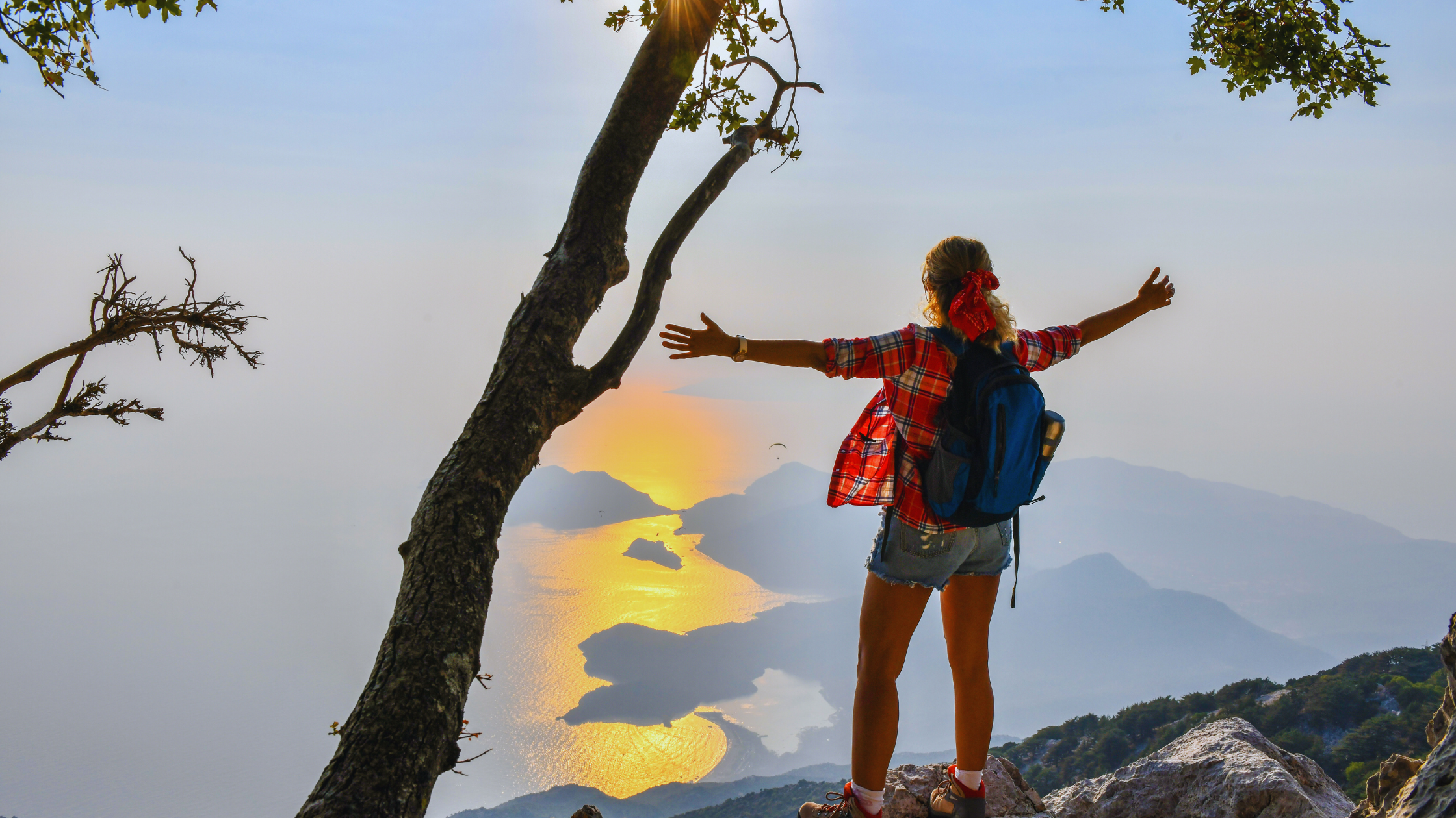 A woman in shorts and a backpack stands on a mountaintop with her arms outstretched. She's facing away from the camera, overlooking a golden river below.