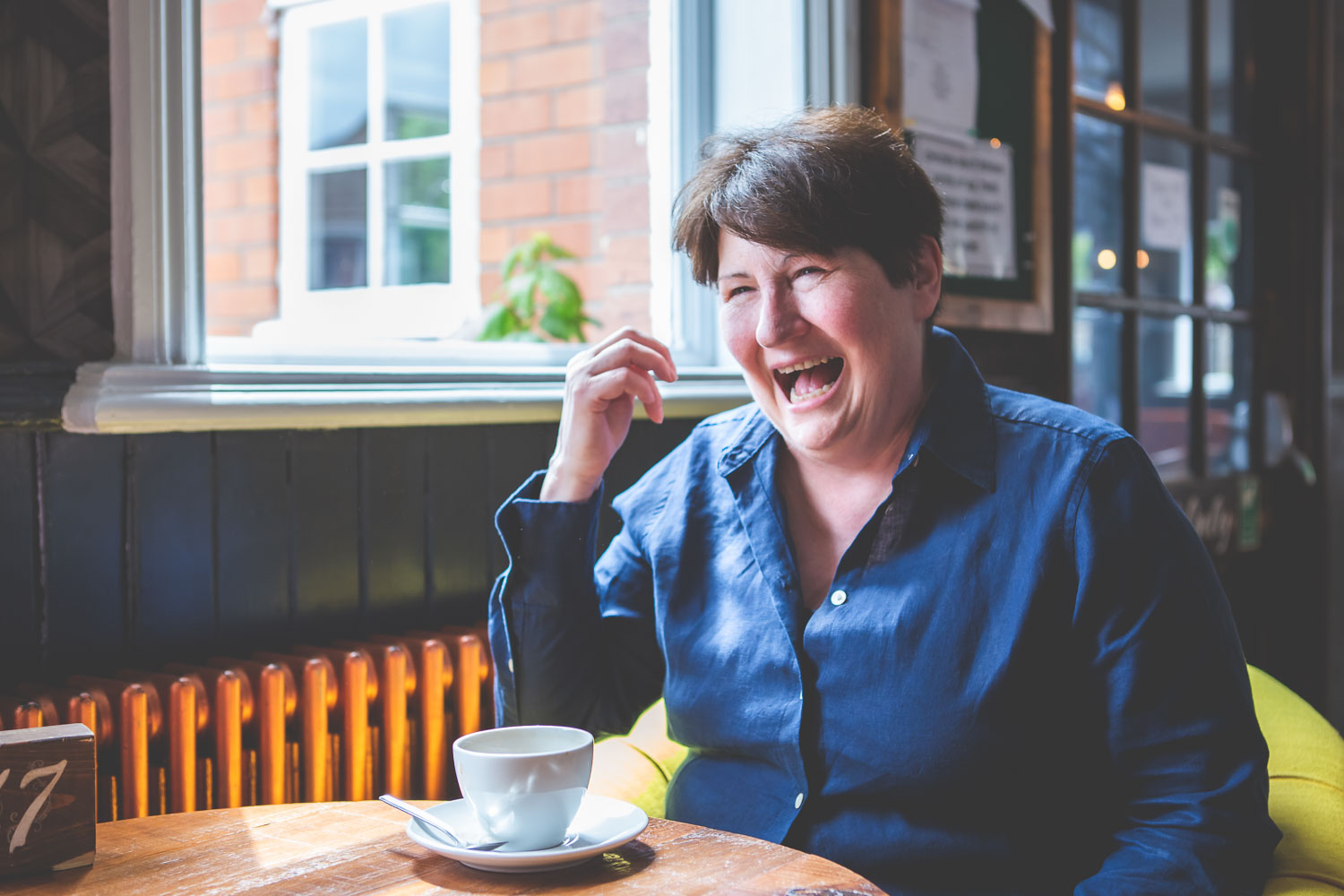 Simone Clarkin, smiling and laughing, enjoys a coffee by a sunny window.