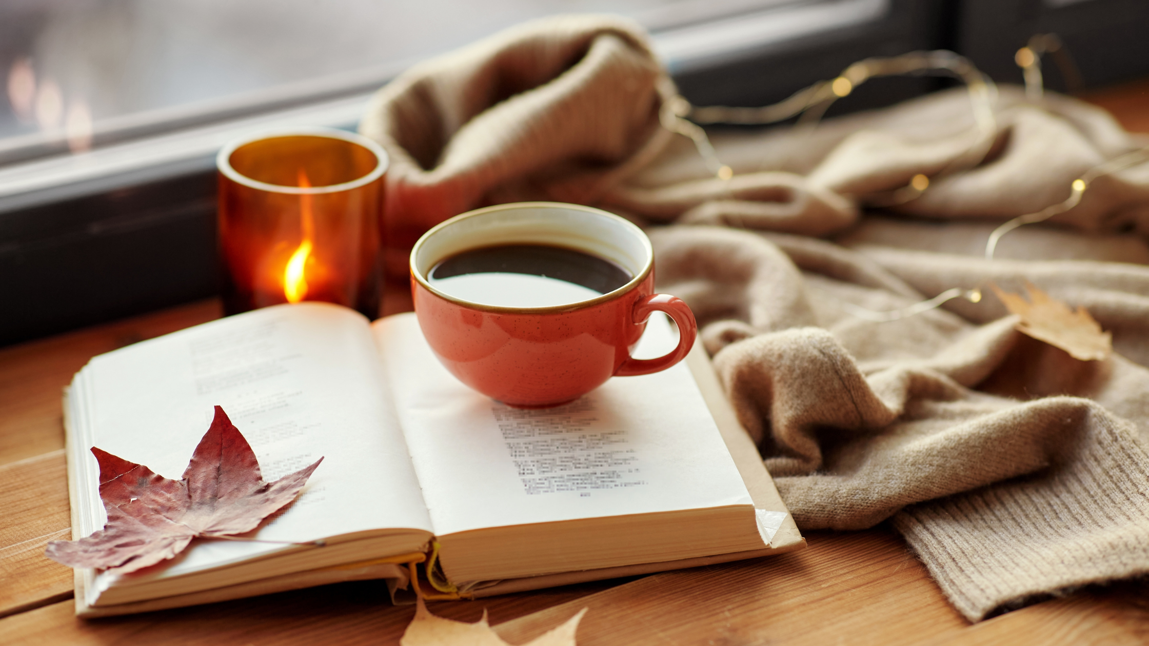 Orange cup of black coffee on open book with autumn leaves by window.