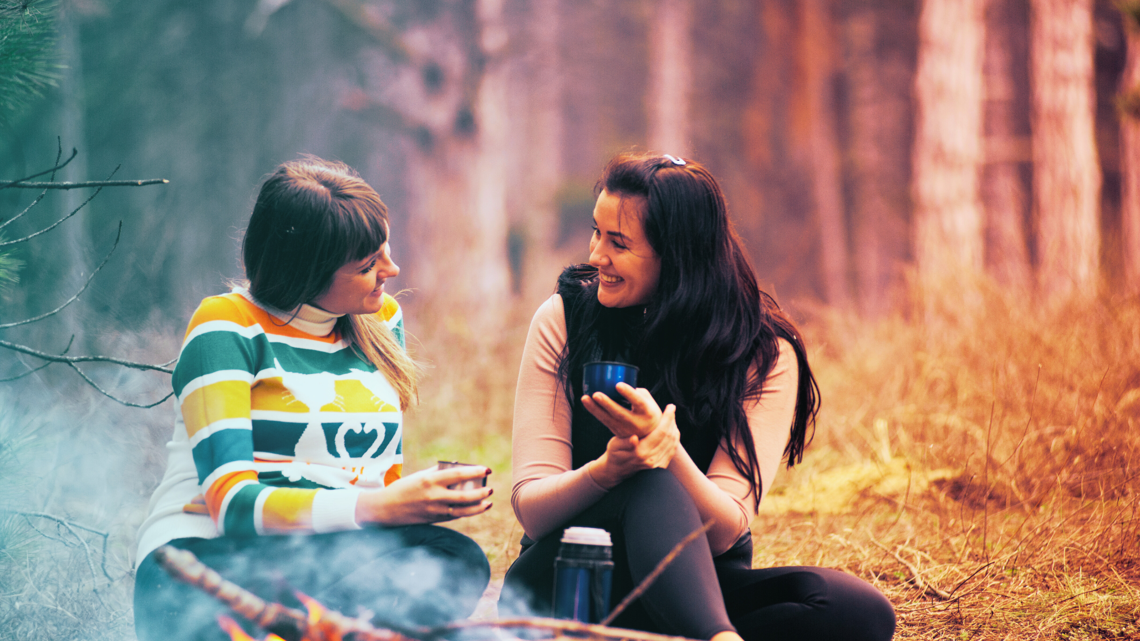 Two young women converse beside an open fire outdoors, sipping a warm beverage.
