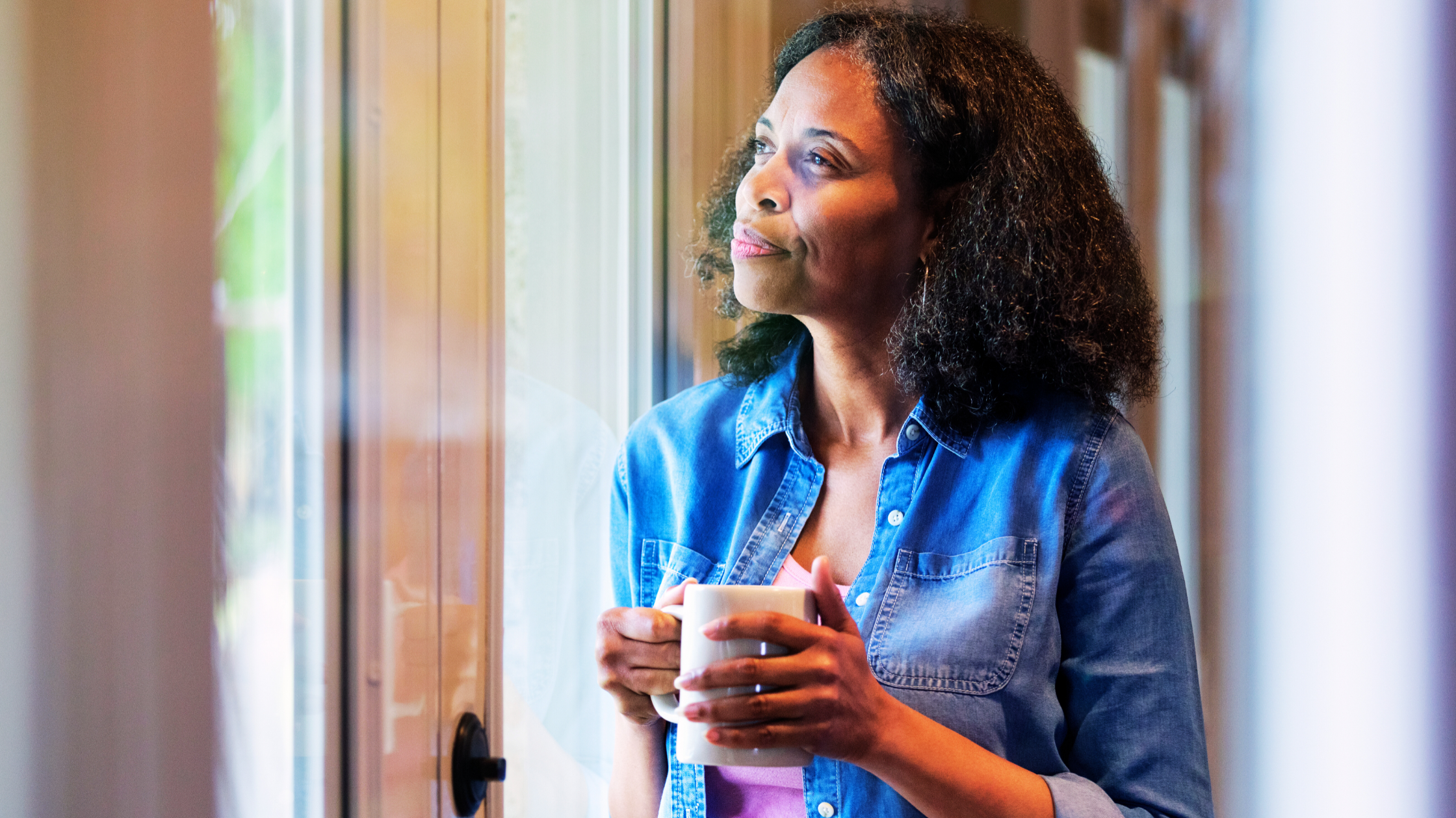 A woman in a blue denim shirt holds a mug and gazes thoughtfully out of a window.
