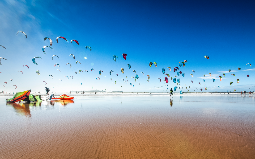 A wide beach with the sea far in the distance, dotted with people flying vibrant, colorful kites under a deep blue sky.
