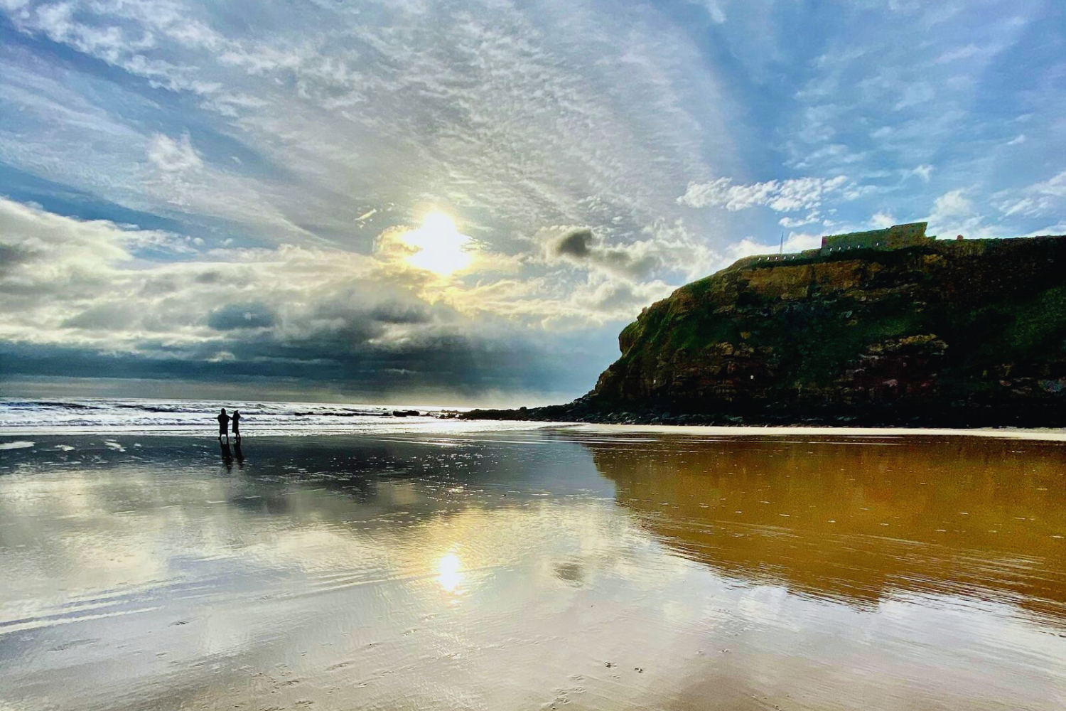 A beach scene with the tide out, revealing wet sand and shallow pools. Low cliffs line part of the shoreline, and a moody, overcast sky looms above. Two people are silhouetted in the distance, walking near the water’s edge.